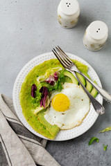 Spinach green crepes (pancakes) with fried egg, avocado and leaves of mix of salad on ceramic plate on gray concrete background. Сoncept of healthy breakfast. Selective focus. Top view. Copt space.