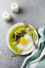 Spinach green crepes (pancakes) with fried egg, avocado and leaves of mix of salad on ceramic plate on gray concrete background. Сoncept of healthy breakfast. Selective focus. Top view. Copt space.