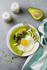 Spinach green crepes (pancakes) with fried egg, avocado and leaves of mix of salad on ceramic plate on gray concrete background. Сoncept of healthy breakfast. Selective focus. Top view. Copt space.