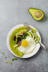 Spinach green crepes (pancakes) with fried egg, avocado and leaves of mix of salad on ceramic plate on gray concrete background. Сoncept of healthy breakfast. Selective focus. Top view. Copt space.