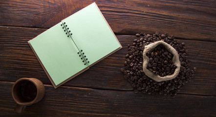 Seeds of coffee, a cup of notebook on a wooden background