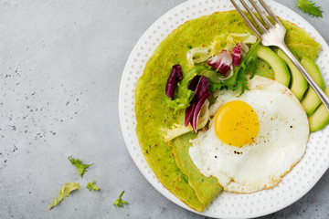 Spinach green crepes (pancakes) with fried egg, avocado and leaves of mix of salad on ceramic plate on gray concrete background. Сoncept of healthy breakfast. Selective focus. Top view. Copt space.
