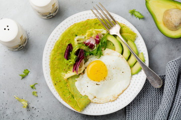 Spinach green crepes (pancakes) with fried egg, avocado and leaves of mix of salad on ceramic plate on gray concrete background. Сoncept of healthy breakfast. Selective focus. Top view. Copt space.