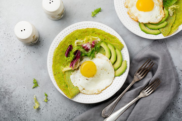 Spinach green crepes (pancakes) with fried egg, avocado and leaves of mix of salad on ceramic plate on gray concrete background. Сoncept of healthy breakfast. Selective focus. Top view. Copt space.