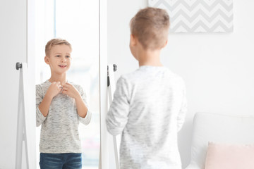 Cute little boy looking at himself in mirror indoors