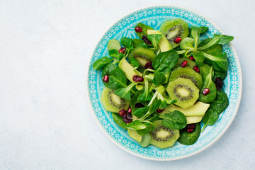 Salad of spinach baby leaves, watercress, kiwi, avocado and pomegranate in blue ceramic plate on white light wooden background. Selective focus. Top view. Copy space.