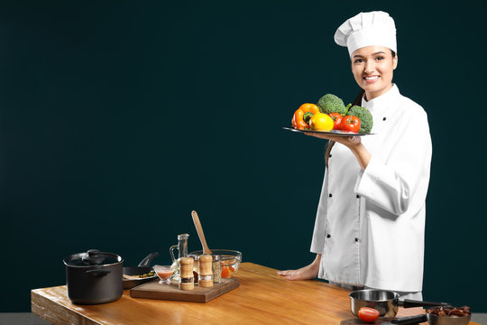 Beautiful female chef holding plate with vegetables near table on color background