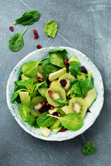 Salad of spinach baby leaves, watercress, kiwi, avocado and pomegranate in old ceramic plate on gray concrete background. Selective focus. Top view. Copy space.