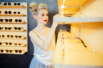 Serious fasionable woman with red lips choosing eyeglasses in optical store - Beautiful girl wearing eyeglasses in optician shop. Ophthalmology concept.