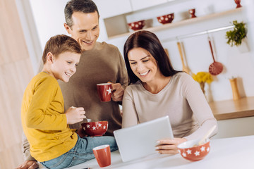 Pleasant breakfast. Upbeat young family having breakfast in the kitchen and watching a video on tablet together while laughing amusingly