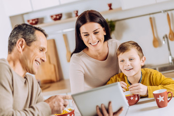 Having fun together. Pleasant young man showing the tablet to his family and watching the video together while they having breakfast in the morning