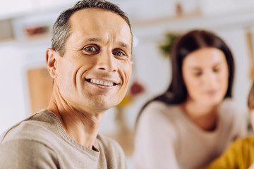 Pleasant smile. The close up of a handsome young man smiling at the camera and posing in the kitchen while his son and wife bonding in the background