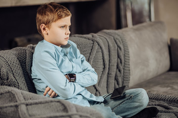 Down in dumps. Cute pre-teen boy sitting cross-legged on the sofa and folding his arms across his chest while looking disappointed, having lost a game