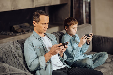 Paying no attention. Pleasant young man and his pre-teen son sitting on the sofa next to each other and being glued to their phones, paying no attention to each other