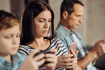 Totally concentrated. Pleasant young woman using her phone and being focused on it while sitting between her husband and son playing games