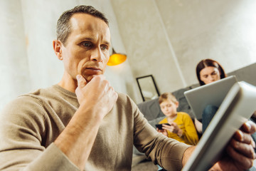 Need to concentrate. Focused young man reading from the tablet and resting his chin on the hand while his family using their devices in the background