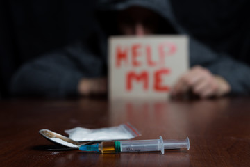 A drug addict at the table holding a sign m asking for help, in front of him a syringe of drugs.