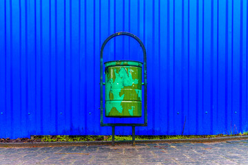 Old green trash can on the background of a blue fence made of metal sheet profile