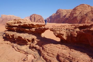 Fototapeta na wymiar Young man standing at natural rock bridge and panoramic view of Jordanian desert Wadi Rum