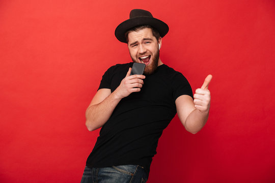 Photo of cheerful man wearing black t-shirt and hat singing while listening to music using mobile phone and wireless earphones, isolated over red wall
