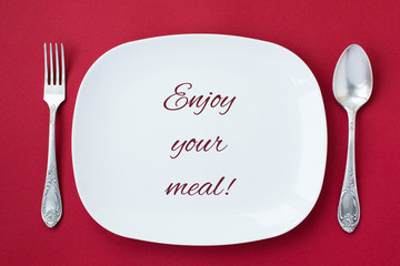 White plate with inscription Enjoy your meal on red background. - obrazy, fototapety, plakaty