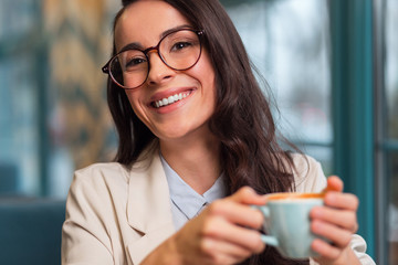 Nice day. Merry positive beautiful woman posing on the blurred background while grinning and carrying cup of coffee