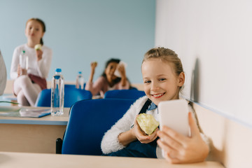 happy teen schoolgirl taking selfie at classroom while eating apple