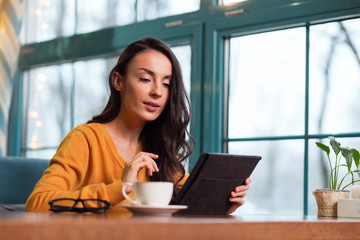 Innovative technology. Musing focused attractive woman sitting at table while using tablet and looking down