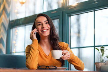 Phone conversation. Gay sincere kind woman getting phone call while smiling near window and sipping tea