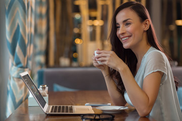 Big opportunity. Satisfied ambitious female freelancer holding cup while sitting at table and using laptop