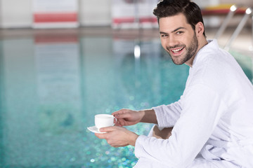 handsome young man in bathrobe holding cup of coffee and smiling at camera in spa center