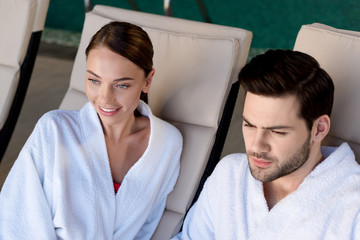 happy young couple in bathrobes smiling and looking away in spa center