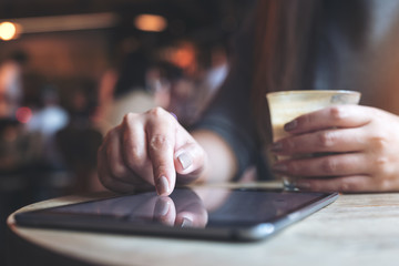 Closeup image of a woman's hand pointing , touching and using tablet pc while holding coffee cup with blur background in cafe