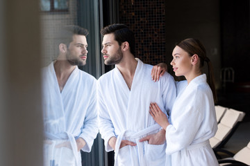 Obraz premium young couple in bathrobes standing together and looking away in spa center