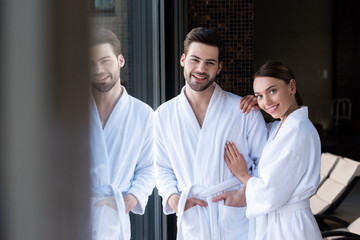 happy young couple in bathrobes smiling at camera in spa center