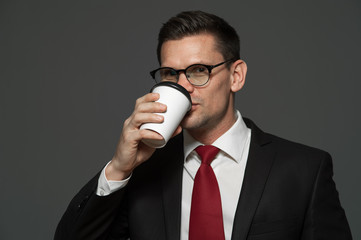 Portrait of young male manager in formal attire and glasses is drinking coffee on gray background