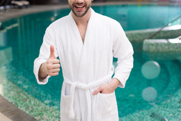 cropped shot of smiling young man in bathrobe showing thumb up in spa center