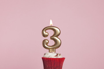 Number 3 gold candle in a cupcake against a pastel pink background