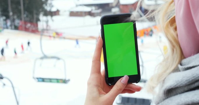close up female hand holding smart phone using mobile tapping scrolling blank green screen chroma key mock-up pointing finger tochscreen blur snow winter skiing people background ski lifts resort