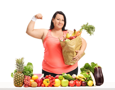 Overweight woman with a paper bag flexing her biceps behind a table with fruit and vegetables