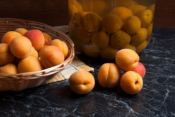 Fresh organic harvested apricots in basket and big glass jar with canned apricot compote on dark marble..