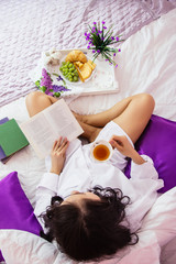Obraz na płótnie Canvas Womans day, morning breakfast white bed, portrait of young brunette girl in pink room Provence style, with purple pillow, book, food, flowers