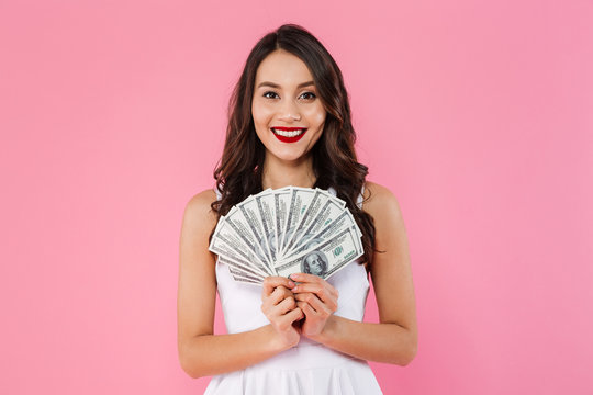 Image of young rich asian woman smiling with white teeth and holding lots of money in dollar currency, isolated over pink background