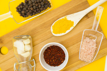 Plastic spoons and cup with dry spices and fresh herbs on a wooden cutting board with yellow background, top view, close up