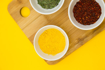 Plastic cups with dry spices and fresh herbs on a wooden cutting board with yellow background, top view, close up
