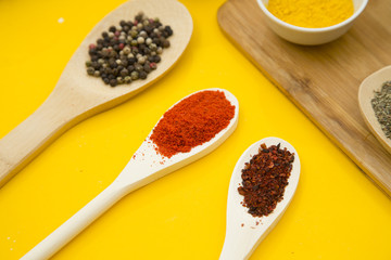 Plastic cups and wooden spoons with dry spices and fresh herbs on a wooden cutting board with yellow background, top view, close up