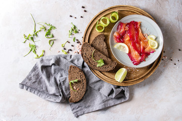 Sliced beetroot marinated salmon for sandwiches with sliced rye bread, pink salt, pepper, greens, cucumber, lemon served on wooden round tray on textile over grey texture background. Top view, space