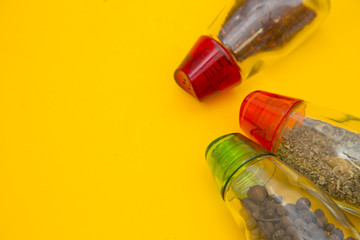Glass bottles with dry spices and fresh herbs on a wooden cutting board with yellow background, top view, close up