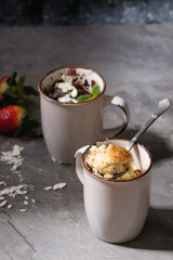 Chocolate and vanilla caramel mug cakes from microwave with fresh strawberries above over grey kitchen table.