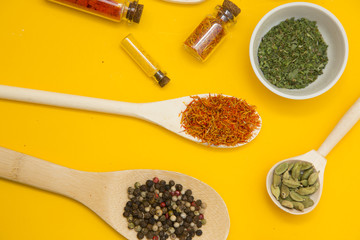 Glass bottles and spoons with dry spices and fresh herbs on a wooden cutting board with yellow background, top view, close up